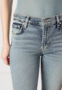 Blugi din denim de culoare bleu deschis, cu un design ajustat, având cinci buzunare, fermoar la brâu și ușoare efecte de uzură aproape de talie.