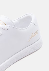 Άσπρο δερμάτινο αθλητικό παπούτσι με υφή στη λευκή σόλα, κορδόνια και χρυσό καλλιγραφικό λογότυπο "Lacoste" στη πλευρά κοντά στη φτέρνα.