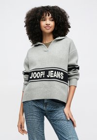Frau mit lockigem Haar, die einen grauen Pullover mit dem Aufdruck "JOOP! JEANS" und blaue Jeans trägt, die mit einem sich wiederholenden "JOOP! JEANS"-Muster bedeckt sind.