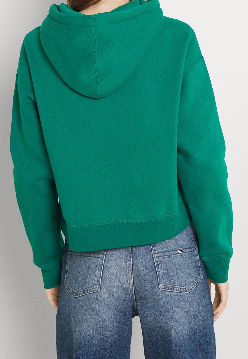 Personne portant un sweat à capuche vert court et un jean en denim bleu, vue de dos sur un fond gris uni.