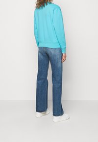 Tyrkysová bavlněná mikina s ribovaným lemem, kombinovaná s modrými rovnými džíny a bílými teniskami. Zobrazení ukazuje zadní část outfitu.