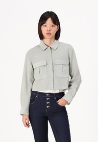 Kort lys grønn jakke med to brystlommer, knappestolpe foran og en myk tekstur, sammen med mørkeblå høytlivsjeans.