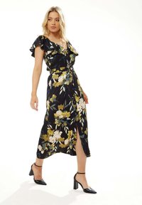 Floral maxi dress in black with yellow and pink flowers, ruffled sleeves, a tie waist, and a front slit, paired with black heels.