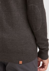 Dunkelgrauer Pullover mit geripptem Saum und Bündchen, ausgestattet mit Ellbogenpatches und einem Lederlogo-Tag in der Nähe des Saums. Der Stoff wirkt glatt und gestrickt.