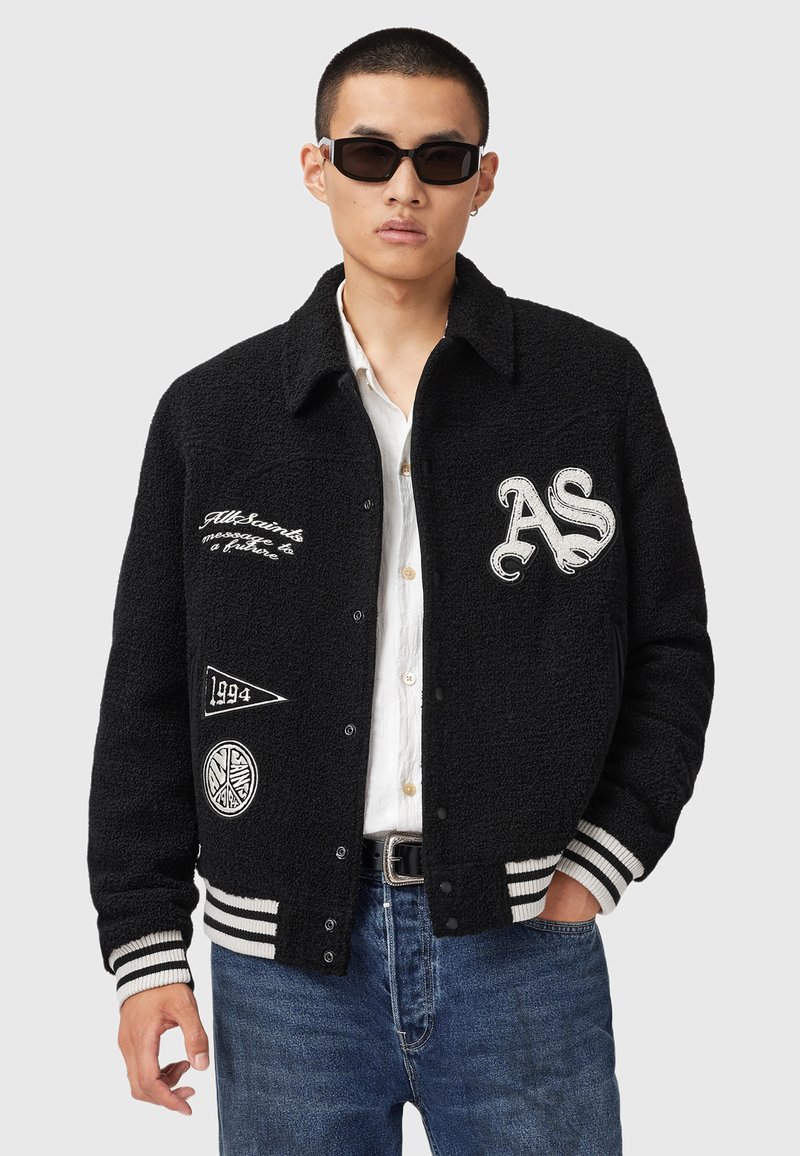 Ung mand iført en sort varsityjakke med hvide lommer, hvid skjorte, blå jeans og sorte solbriller, poserer med den ene hånd i lommen.