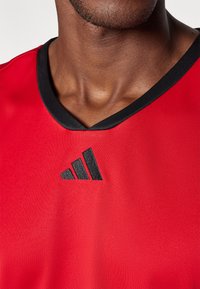 Camiseta atlética roja con cuello en V negro y un logo bordado negro que presenta tres franjas verticales en el lado izquierdo del pecho. Textura suave.