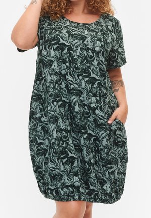 Frau trägt ein kurzärmeliges, knielanges, grünes Kleid mit schwarzem abstraktem Blumenmuster und elastischem Saum, Hand in der Tasche.