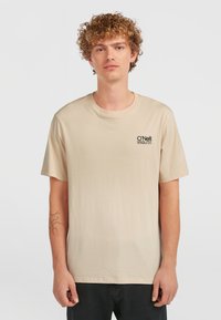 T-shirt in cotone beige con maniche corte, collo a giro, e logo nero sul petto sinistro. Il modello è in piedi davanti a uno sfondo neutro.