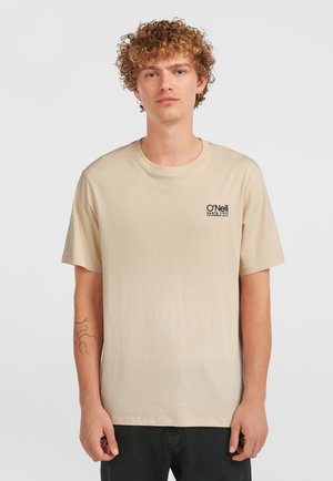 Beige Baumwoll-T-Shirt mit kurzen Ärmeln, Rundhalsausschnitt und schwarzem Logo auf der linken Brust. Modell steht vor einem einfarbigen Hintergrund.