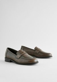 Loafers din piele maro cu un finisaj neted, având o curea decorativă în față și un toc solid, de înălțime mică. Design minimalist.