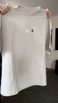 Camiseta blanca sostenida por manos, con pequeño texto "Pegador" y un gráfico de mariposa morada cerca de la zona del pecho.