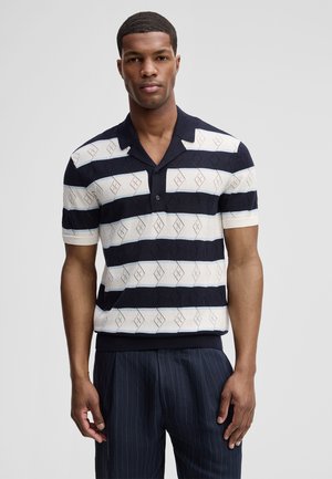 Mann trägt ein navy-weiß gestreiftes kurzärmeliges Strickpolo mit Rautenmuster, kombiniert mit dunkler Nadelstreifenhose, steht vor weißem Hintergrund.