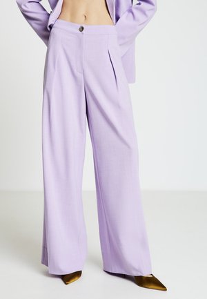 Pantalon larges couleur lilas avec taille haute, plis et un bouton décoratif. Texture de tissu lisse, associé à des talons pointus dorés.