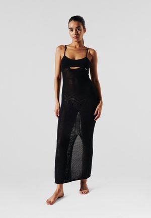 Black, textured knit maxi dress with spaghetti straps and a front cutout; features a form-fitting silhouette. Barefoot model stands against a plain background.