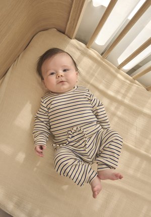 Body a righe beige e blu navy per neonati, con maniche lunghe e dettaglio annodato, posato su un materasso per culla di colore neutro chiaro con una struttura di legno.