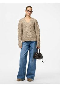 Beige gebreide trui met een V-hals en een textuur kabelpatroon, gecombineerd met wijde blauwe jeans en een kleine zwarte handtas.