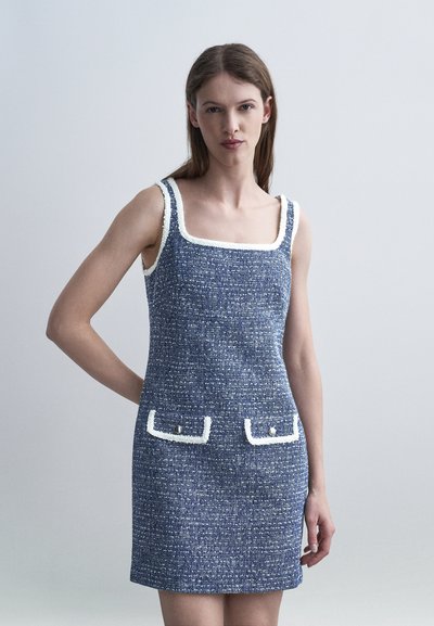 Femme portant une robe bleue texturée sans manches avec bordure blanche et deux poches avant à boutons, debout devant un fond clair uni.
