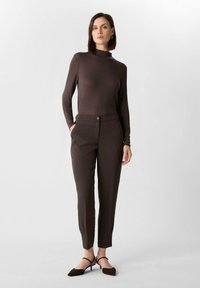 Top lungo marrone a maniche lunghe con scollo alto e silhouette aderente, abbinato a pantaloni marroni su misura con bottone frontale e tasche inclinati.
