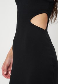 Gros plan d’une robe noire avec une découpe latérale laissant apparaître la peau, et une main reposant près de la hanche.