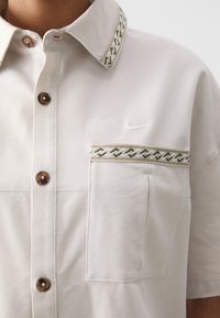 Lys beige skjorte med krage som har mønstrede detaljer, frontlomme og brune knapper. Inkluderer en brodert Nike-logo på brystet.