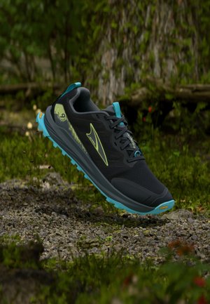 Chaussure de trail running noire et bleue avec logo jaune suspendue au-dessus d'un sol rocheux en forêt, avec des arbres et de la verdure en arrière-plan.