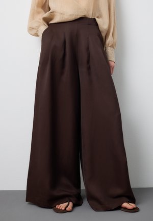 Blusa beige de manga larga metida en pantalones anchos de tiro alto color marrón oscuro, combinados con sandalias marrones sobre un suelo gris.