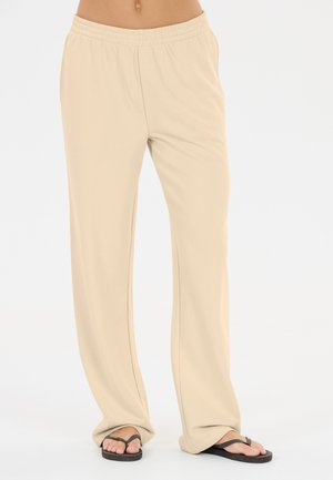 Beige løsere bukser med elastik i taljen, båret med sorte flip-flops, vist fra talje til fødder på en lys baggrund.