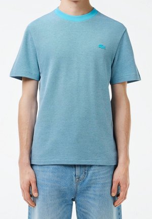 Homme portant un t-shirt texturé bleu clair à manches courtes avec col turquoise et petit logo brodé sur la poitrine, associé à un jean bleu clair.