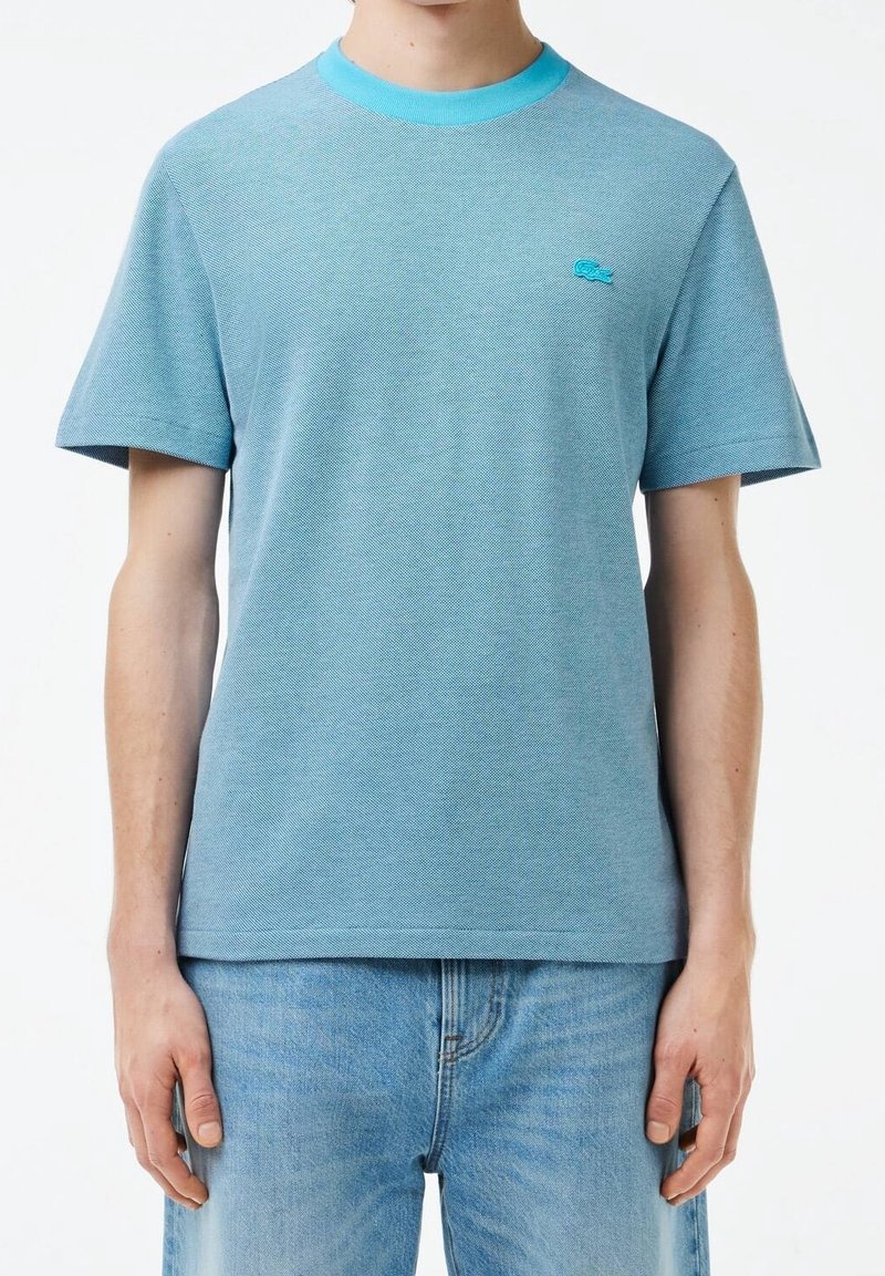 Homme portant un t-shirt texturé bleu clair à manches courtes avec col turquoise et petit logo brodé sur la poitrine, associé à un jean bleu clair.