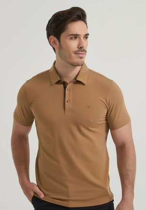 Homme aux cheveux courts et foncés portant un polo marron à manches courtes, se tenant devant un fond clair uni.