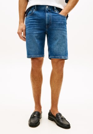 Mand iført blå denimshorts med forlommer, en hvid skjorte og sorte læder slip-on loafers, stående med den ene hånd i lommen.