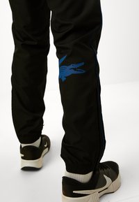 Pantaloni sport negri cu un logo al unui crocodil albastru pe genunchiul stâng, având dungi laterale albastre și o margine tivită. Purtați cu adidași negri.