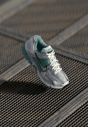 Chaussure de running Nike blanche et turquoise avec logo Swoosh noir flottant au-dessus d’une surface en grille métallique texturée.