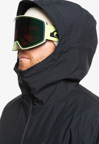 Schwarze Jacke mit Kapuze, strukturiertem Stoff und Reißverschluss. Getragen mit grünen Skibrillen und einer leichten Strickmütze darunter.