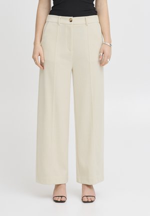 Persoon draagt beige wijde broek, zwarte mouwloze top, zwarte sandalen en zilveren sieraden, staand tegen een effen witte achtergrond.