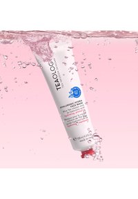 Tubo bianco con tappo rosso etichettato "Maschera Viso Idraulica Idratante". Testo in blu e rosa. Sott'acqua chiara con bolle, sfondo rosa.