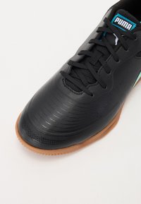 Zapatilla deportiva negra con parte superior sintética texturizada, cordones negros, acentos en teal y suela de goma. Presenta líneas horizontales en la puntera.