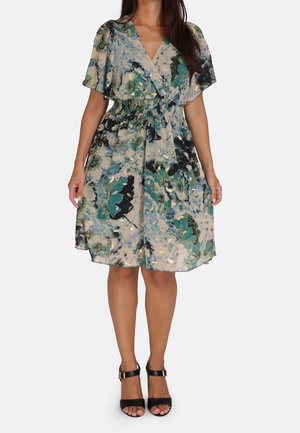 Une femme porte une robe courte aux genoux, à manches courtes, avec un motif floral vert océan et beige, et des sandales noires à talons hauts avec bride à la cheville.