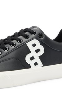 Nära bild av en svart sneakers med svarta snörningar och vit sula, prydd med en broderad vit dubbel-B-logotyp på sidopanelen.