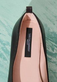Gros plan d'une chaussure plate en cuir noir avec intérieur rose clair et une étiquette noire indiquant « Tabitha Simmons Next » sur une surface verte texturée.