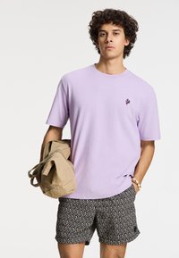 Lavender spalvos medvilnės marškinėliai su apvalia iškirpte ir mažai tamsia logotipu, derinami su raštuotais juodais šortais. Modelis laiko kreminės spalvos maišelį.