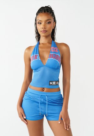 Kobieta w niebieskim sportowym topie halter i pasujących szortach, stojąca na białym tle.