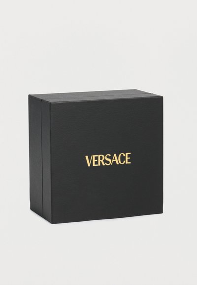 Boîte carrée noire avec logo VERSACE doré centré sur le devant, sur un fond clair uni.