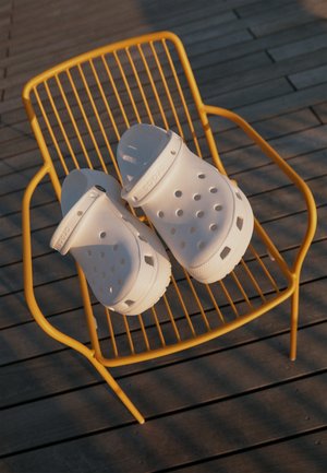 Sabots blancs à enfiler avec des trous de ventilation posés sur une chaise en fil métallique jaune sur une terrasse en bois sous une lumière douce du soleil.