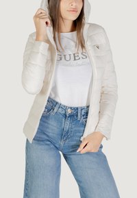Witte pufferjack met een capuchon, met een zachte textuur, ritsluiting en een klein logo-detail. Draag het over een lichtgekleurde casual shirt en denim jeans.