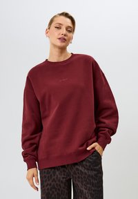 Bordeaux sweatshirt met een ronde hals, relaxte pasvorm en ribgebreide manchetten. Bevat subtiele branding op de borst. Gecombineerd met patroonsbroek.
