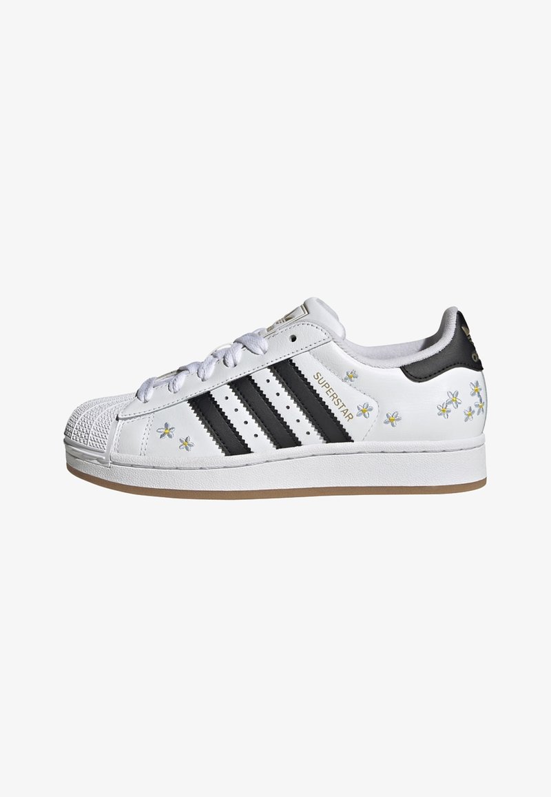 Valkoinen Adidas Superstar -tennari mustilla raidoilla, pienillä keltaisilla ja valkoisilla kukkakuoseilla ja kumipohjalla, sivulta katsottuna.