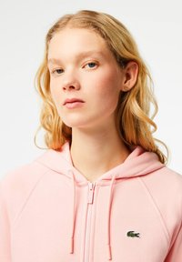 Plavuša koja nosi ružičastu duksericu na zip s Lacoste logom i malim zelenim minđušama u obliku krokodila, samouvjereno gleda u kameru.