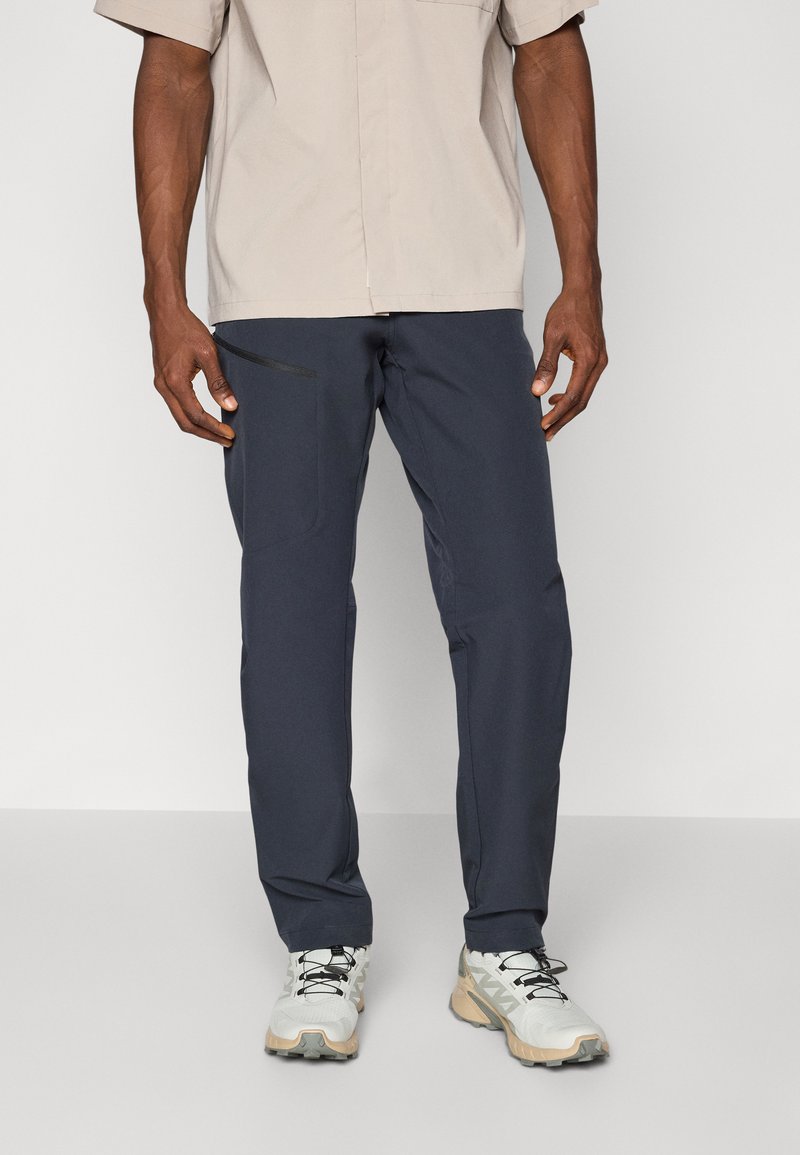 Mørkeblå, letvægtsbukser med slim fit, med en sidelomme med en sort lynlås, parret med hvide og beige træningssko.