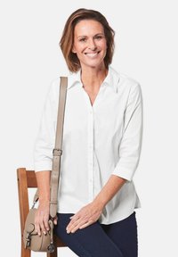 Camicia bianca con bottoni e maniche lunghe. Slim fit, tessuto morbido, colletto ampio. Indossata con pantaloni blu scuro e una borsa a tracolla beige.